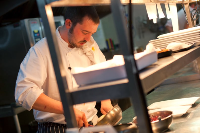 A chef stood at the pass plating a dish up with further orders on the rack