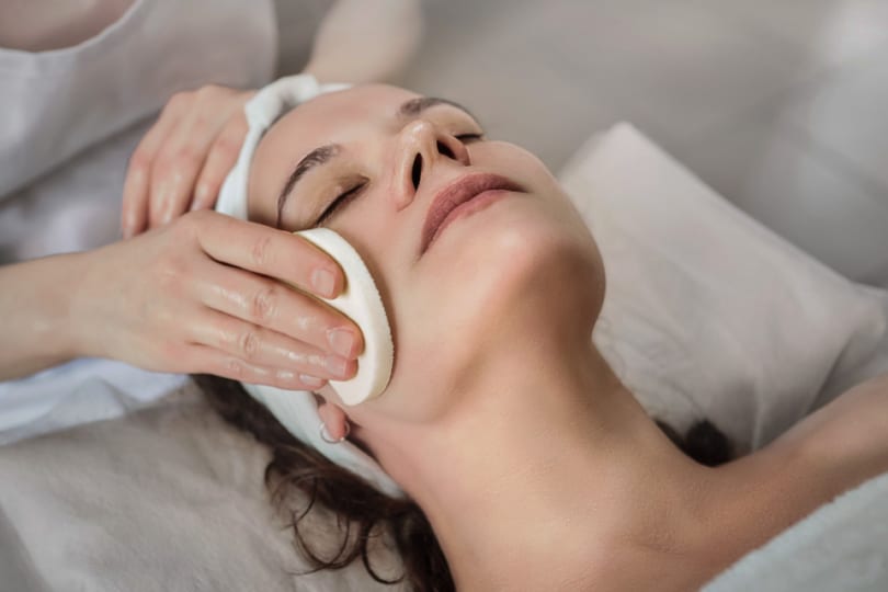 A lady lying on her back with her eyes closed receiving a facial treatment by a spa therapist